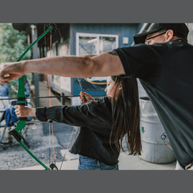 Archery Lessons with an instructor showing a kid that has a bow and arrow ready to shoot
